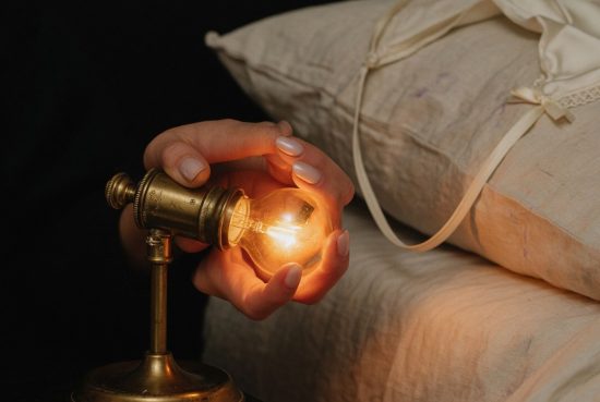 Gros plan d'une main allumant une lampe de chevet vintage, draps en lin froissés en arrière-plan, chambre dans la pénombre