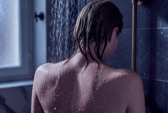 Femme vue de dos sous douche froide, cheveux blonds mouillés, gros plan nuque et épaules, gouttes d'eau.