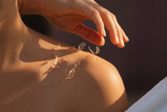 Épaule en lumière dorée, fine peau qui pèle, main dont les doigts effleurent la surface.