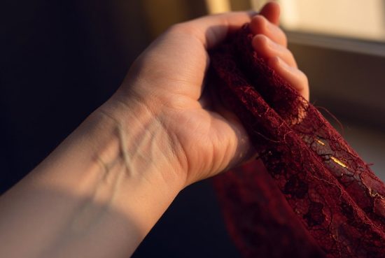 Gros plan vertical d'un poignet féminin, veines apparentes sous lumière dorée, dentelle rouge grenat effleurant le cadre.