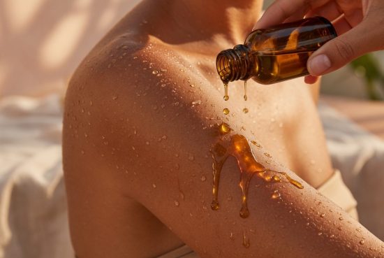Gros plan d'une épaule bronzée couverte de gouttes d'eau, une main versant de l'huile dorée d'une fiole ambrée sur la peau.