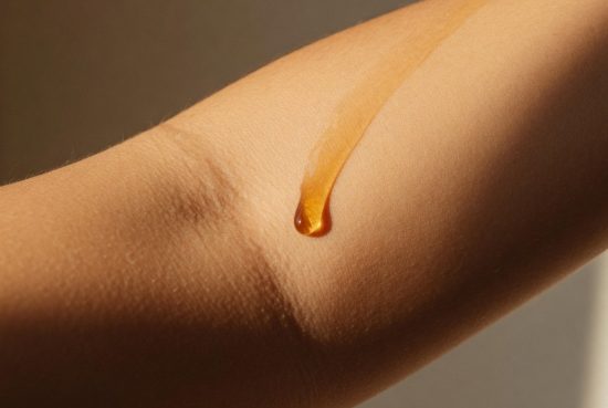 Gros plan du coude en lumière rasante, texture naturelle de la peau avec une goutte de sérum brillante.