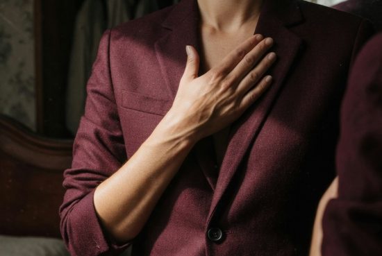 Femme en blazer bordeaux, main au revers, épaule nue, face hors champ devant miroir ancien.